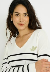 Suéter de cuello en V color crema con rayas horizontales negras. Presenta un bordado de corazón verde en el pecho. Textura de punto suave, ajuste regular.