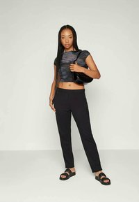 Zwart cropped top van textuurstof, strakke zwarte broek en zwarte sandalen. Model houdt een zwarte schoudertas vast en heeft lange vlechten.