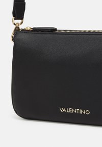 Svart läder crossbody-väska med texturerad yta, guldfärgad hårdvara och dragkedja. Har det präglade "Valentino"-logotypen i guld på framsidan.