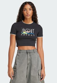 Černé zkrácené tričko s bílým lemem a barevným logem Adidas. V kombinaci s šedými cargo kalhotami s vysokým pasem a bočními kapsami.