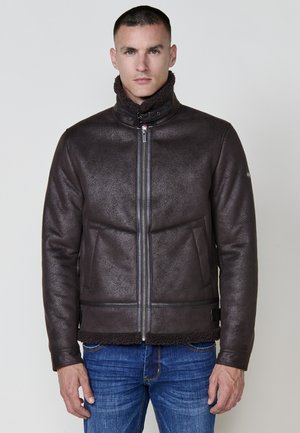 Chaqueta de cuero sintético marrón con un acabado texturizado, cierre de cremallera, cuello alto y forro de piel sintética en la parte inferior, combinada con jeans azules.