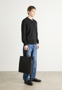 Schwarze Tote-Bag mit zwei stabilen Griffen, rechteckiger Form und glatter Textur, kombiniert mit einem schwarzen Pullover und blauen Jeans.