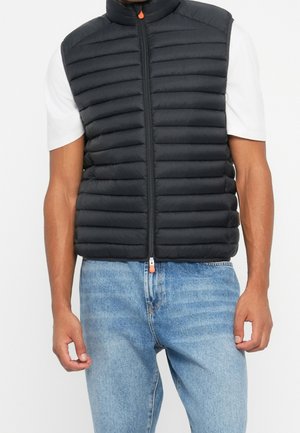 Mężczyzna ubrany w czarną pikowaną kamizelkę bez rękawów na białą koszulę z krótkim rękawem i jasnoniebieskie dżinsy, stoi z rozluźnionymi ramionami.