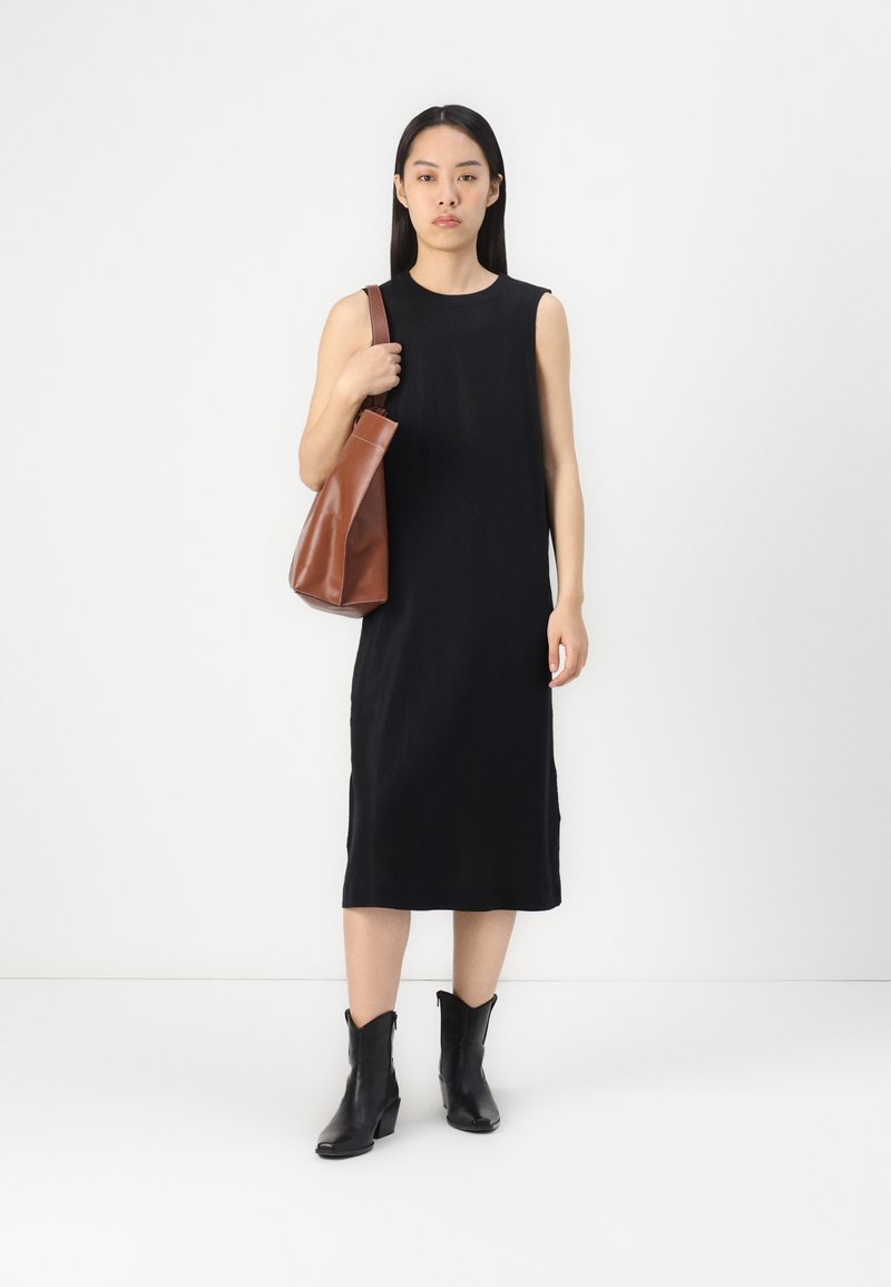 Schwarzes ärmelloses Kleid mit geradem Schnitt und weicher Textur. Kombiniert mit einer braunen Ledertasche und schwarzen Stiefeln im Western-Stil.