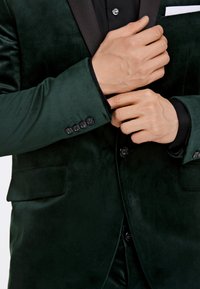Blazer in velluto verde scuro con colletto in satin nero, quattro bottoni neri sui polsini e taschino sul petto con un fazzoletto bianco.