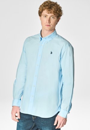 Giovane uomo con capelli ricci che indossa una camicia azzurra chiara con bottoni e un piccolo logo scuro sul petto a sinistra e jeans scuri.