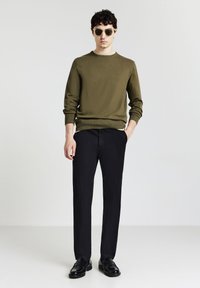 Olivgrüner Strickpullover, schwarze Hose und schwarze Schuhe. Das Model steht aufrecht und trägt Sonnenbrillen. Glatte Textur, runder Halsausschnitt.