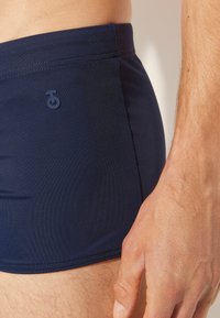Marineblaue Badeshorts aus glattem Stoff, mit einem kleinen Logo an der Seite und flachen Nähten für zusätzlichen Komfort. Nahaufnahme von Passform und Textur.