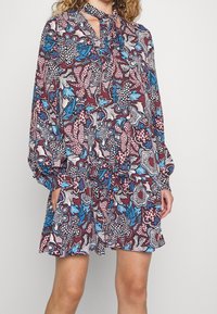 Vícebarevné šaty se květinovým a abstraktním vzorem v burgundské, modré a bílé barvě. Dlouhé rukávy s detailem na zavázání u krku a nařaseným pasem.