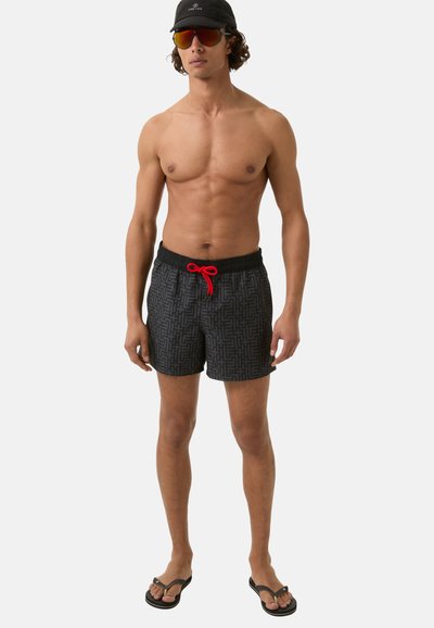 Zwarte zwemshorts met een geometrisch patroon, rode trekkoord en een elastische tailleband. Vergezeld van slippers en een zonnebril met spiegels.