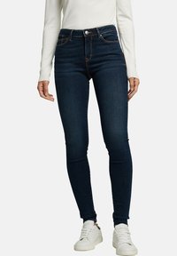 Jeans de tiro alto en azul oscuro con un diseño ajustado y acentos sutiles, hechos de denim. Combinados con zapatillas blancas y un suéter ligero.