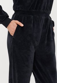 Tuta nera in velluto con vita elastica, maniche lunghe e tasche laterali, caratterizzata da una consistenza morbida e liscia in tutto il suo insieme.