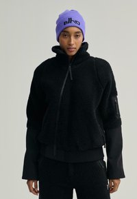 Černá fleecová bunda s vysokým límcem, zipovým uzávěrem a žebrovanými manžetami. V kombinaci s černými fleecovými kalhotami. Fialová pletená čepice s logem.