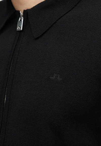 Κοντινό πλάνο μαύρου πλεκτού πουλόβερ με φερμουάρ, γιακά και διακριτικό κεντημένο λογότυπο "JL" στο στήθος.