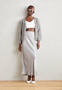 O femeie îmbrăcată cu un top alb pe burtă, un cardigan fuzzy gri, o fustă satinată argintie, tocuri albe, ochelari de soare albi și bijuterii aurii, stând lângă un perete simplu.