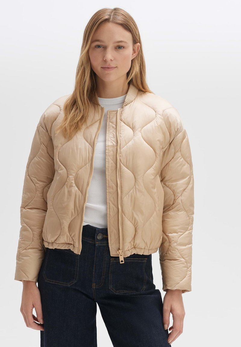 Beige gesteppte Bomberjacke mit geripptem Kragen und Reißverschluss. Der strukturierte, glänzende Stoff kontrastiert mit dunklen Denim-Hosen.