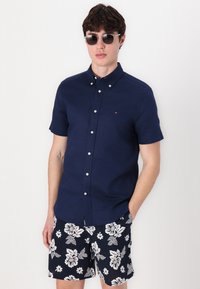 Marineblå kortermet skjorte med knapper kombinert med shorts med svart-hvitt blomstertrykk. Skjorten har klassisk krage og hvite knapper.