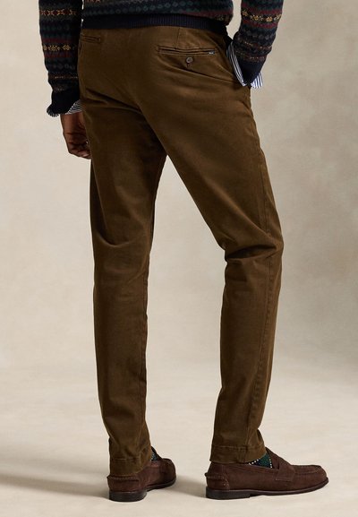 Άνδρας που φοράει καφέ παντελόνια slim-fit, σκουροκαφέ μοκασίνια και πουλόβερ με σχέδιο, στεκόμενος με τα χέρια στις τσέπες, εμφανίζεται από πίσω.