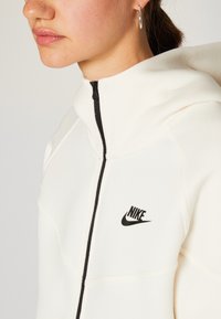 Nike mikina v krémové barvě, se černým zipem a logem. Design zahrnuje vysoký límec a prošívané detaily pro texturu.