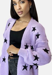 Fioletowy, dziergany kardigan o luźnym kroju, z długimi rękawami i czarnymi wzorami w gwiazdki. Połączony z czarnym topem i ciemnymi dżinsami.
