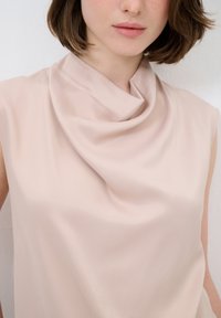 Top senza maniche con collo a cowl, realizzato in morbido tessuto rosa pallido, caratterizzato da delicate pieghe e una silhouette aderente.