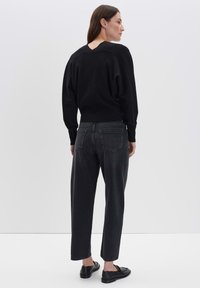 Eine Frau steht und trägt ein schwarzes Langarmoberteil, hochgeschnittene schwarze Jeans und schwarze Slipper. Sie steht seitlich und hat den Kopf nach rechts gedreht.