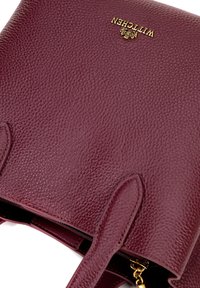 Bordeaux lederen handtas met een gestructureerde afwerking, voorzien van gouden hardware en logo-detail. Inclusief dubbele handvatten en ritssluiting.
