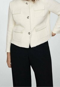 White tweed jacket with a boxy cut, four front pockets, and silver buttons, paired with black wide-leg trousers. Soft texture and frayed edges.