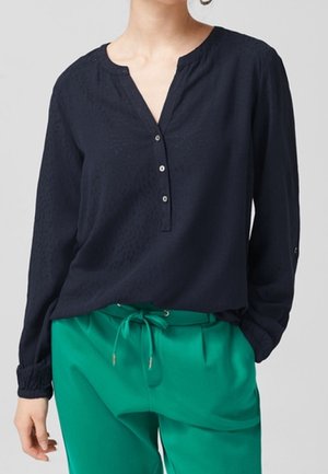 Femme portant un chemisier bleu marine à manches longues avec des boutons et un pantalon vert à cordon, posant avec une main dans la poche.
