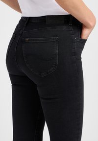 Jean en denim noir au design ajusté, avec une poche arrière et une ceinture texturée. Comprend un petit accent logo doré sur la poche.