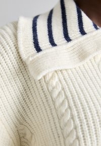 Cremefarbener, gestrickter Pullover mit strukturiertem Körper, verfügt über einen gerippten Kragen und drei marineblaue Streifen am Kragen. Zopfmuster-Detail an der Seite.