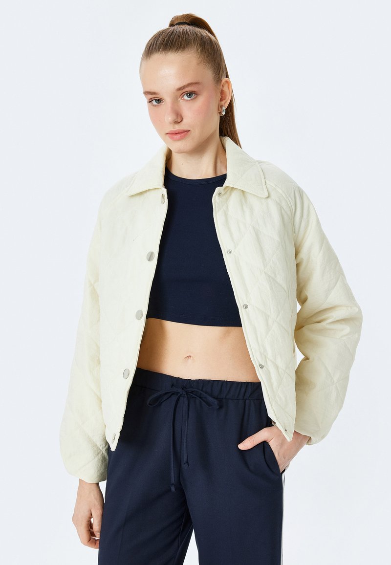Gepolsterte cremefarbene Jacke mit Kragen, Druckknöpfen und langen Ärmeln, getragen über einem marineblauen Cropped-Top. Glatte Textur, lässige Passform.