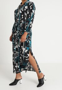 Lång svart klänning med vita och teal blommönster, med krage, långärmade och sidoklyftor. Matchas med svarta klackmules.