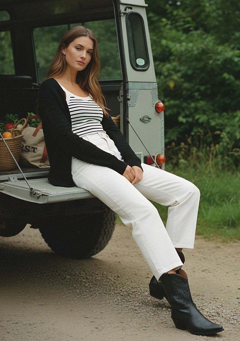 Black cardigan, striped tank top, high-waisted white pants, and black cowboy boots. Sitting on a truck tailgate, green background.