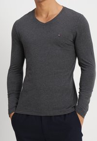 Mann trägt ein enganliegendes, dunkelgraues Langarmshirt mit V-Ausschnitt und einem kleinen rot-weißen und blauen Logo auf der linken Brust.