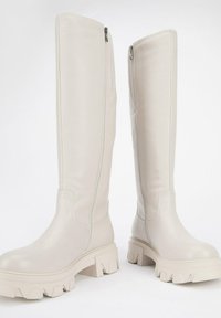 Botas de cuero suave de color beige claro, hasta la rodilla, con cremalleras laterales y suelas gruesas y texturizadas con un diseño robusto.