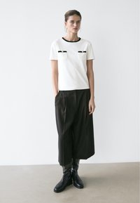 Femme debout contre un mur blanc, portant un haut blanc à manches courtes avec bordure noire, un pantalon large noir coupé au-dessus de la cheville et des bottes noires montant jusqu'aux genoux.