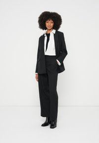 Kvinne står med hånden i lommen, iført svart blazer, svarte vide bukser, svarte støvler, hvit skjorte og svart topp mot en ensfarget hvit bakgrunn.