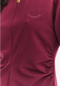 Bordowy krótki rękaw bawełniana bluzka z kołnierzykiem na guziki, ozdobiona różowym haftowanym motywem rzęs oraz marszczeniami po bokach.