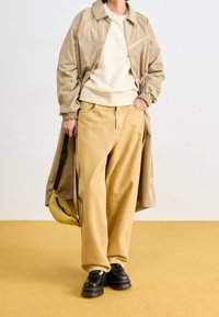 Béžový trenčkot přes světlou mikinu, doplněný širokými béžovými kalhotami a černými robustními botami. Z jednoho ramene visí malá žlutá taška.