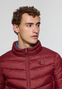 Chaqueta acolchada burdeos con un alto cuello, cierre de cremallera y un parche de logo en el lado izquierdo del pecho. Presenta una textura acolchada y un diseño ligero.