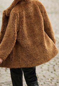 Chaqueta marrón de piel sintética con un exterior texturizado y esponjoso, y un ajuste relajado. Presenta un cuello tipo chal y se combina con pantalones negros ajustados.