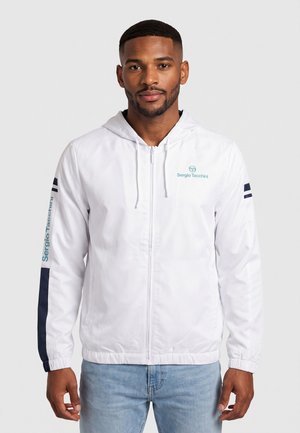 Hombre que lleva una sudadera con capucha blanca con cremallera, una franja azul marino en la manga izquierda y el logo de Sergio Tacchini en el pecho y la manga.