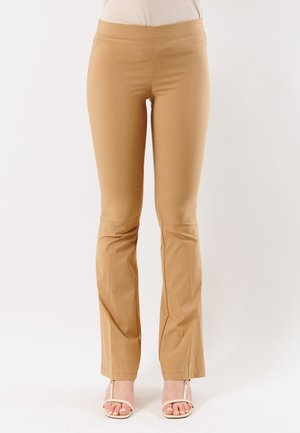 Donna che indossa pantaloni aderenti beige abbinati a sandali crema con punta aperta, in piedi contro uno sfondo chiaro e semplice.