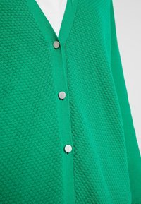 Zelený kardigan s texturovaným pleteným vzorem, s výstřihem do V a třemi kovovými knoflíky vpředu. Měkká látka a přiléhavý střih.