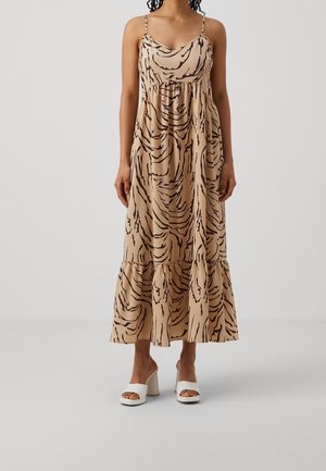 Vrouw draagt een beige midi-jurk met bruine abstracte draaipatronen, spaghetti bandjes en witte sandalen met hoge hakken, staand tegen een effen achtergrond.