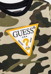 Tă fabrică cu model de camuflaj în nuanțe de verde și bej, având un patch triunghiular galben cu logo-ul "GUESS" și detalii de text. Guler negru cu caneluri.