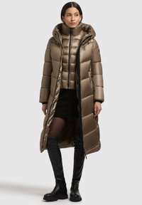Long manteau en duvet vert olive avec capuche, présentant une texture brillante, un design matelassé et des accents de fermeture éclair noirs. Porté par-dessus une veste courte.