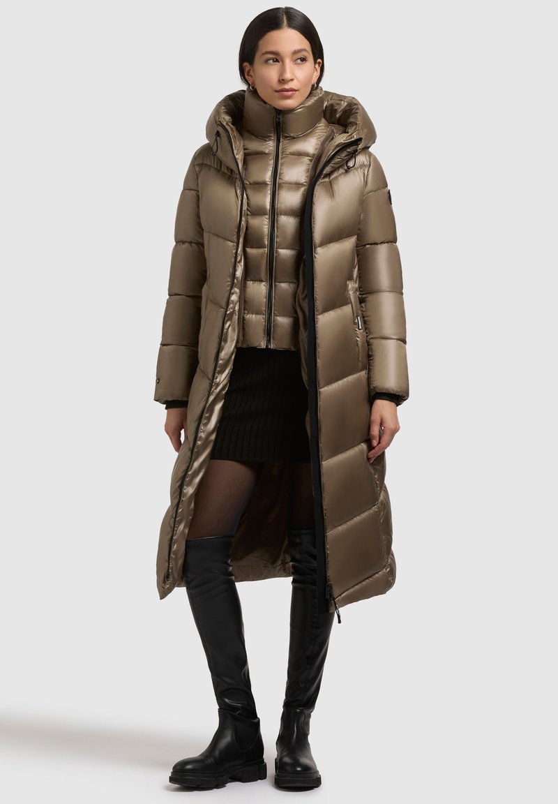 Long manteau en duvet vert olive avec capuche, présentant une texture brillante, un design matelassé et des accents de fermeture éclair noirs. Porté par-dessus une veste courte.