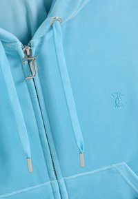 Svetlomodrá mikina s kapucňou a sťahovacími šnúrkami, so strieborným zatváraním na zips v tvare písmena „J“ a malým logom z trblietavých kamienkov na hrudi.